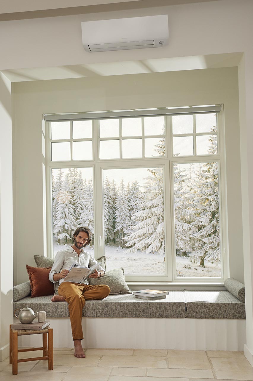 Mies istuu ikkunalaudalla lukemassa lumisen metsänäkymän edessä, modernissa olohuoneessa.